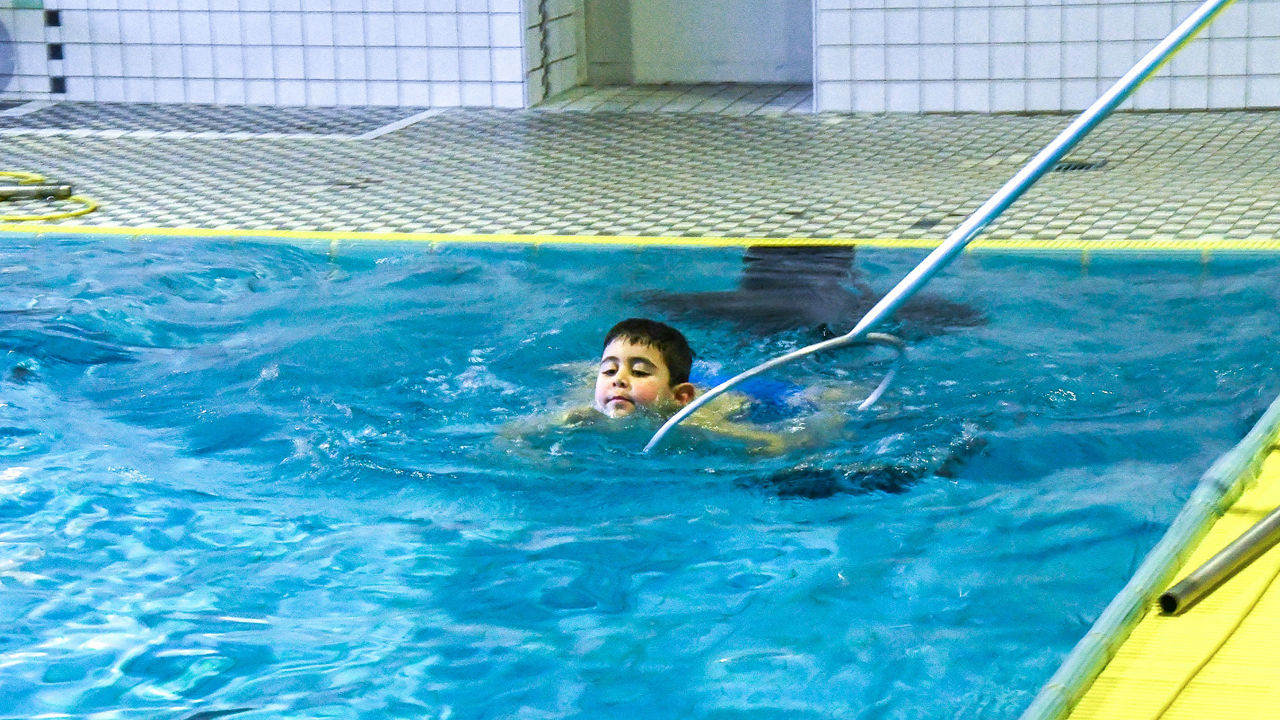 Ein Junge schwimmt im Schwimmer-Bereich kurz hinter dem Beckenrand. Ein Betreuer reicht ihm einen Stab mit Ring als Hilfe.
