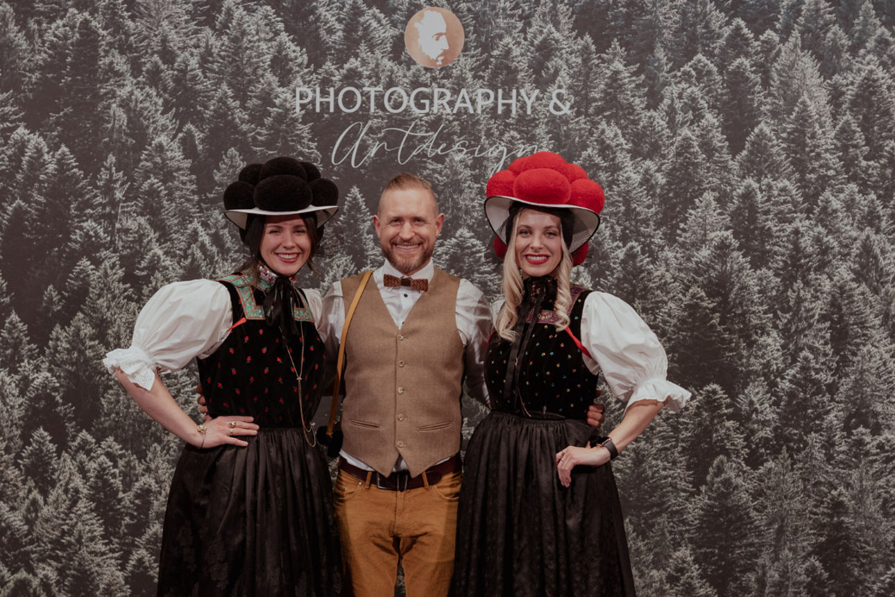 Fotokunst von Sebastian Wehrle: Highlandrind mit Blumenkranz neben Person in Tracht mit rotem Bollenhut.