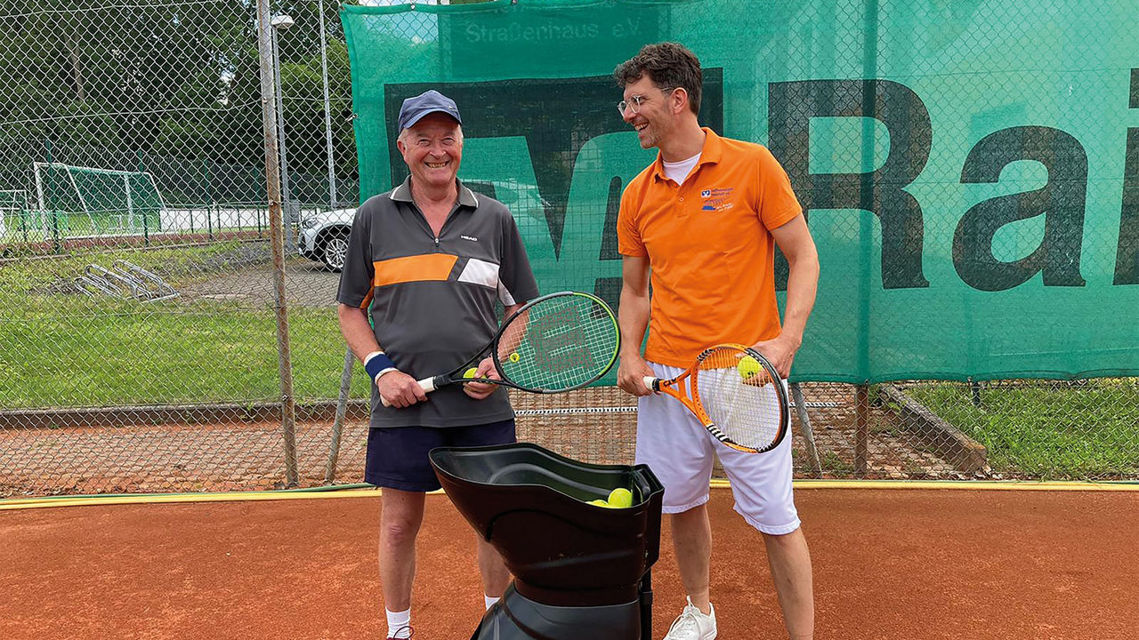 Roland Pees und ein Mitglied des Tennisclubs Straßenhaus stehen mit Tennisschlägern neben der Ballmaschine