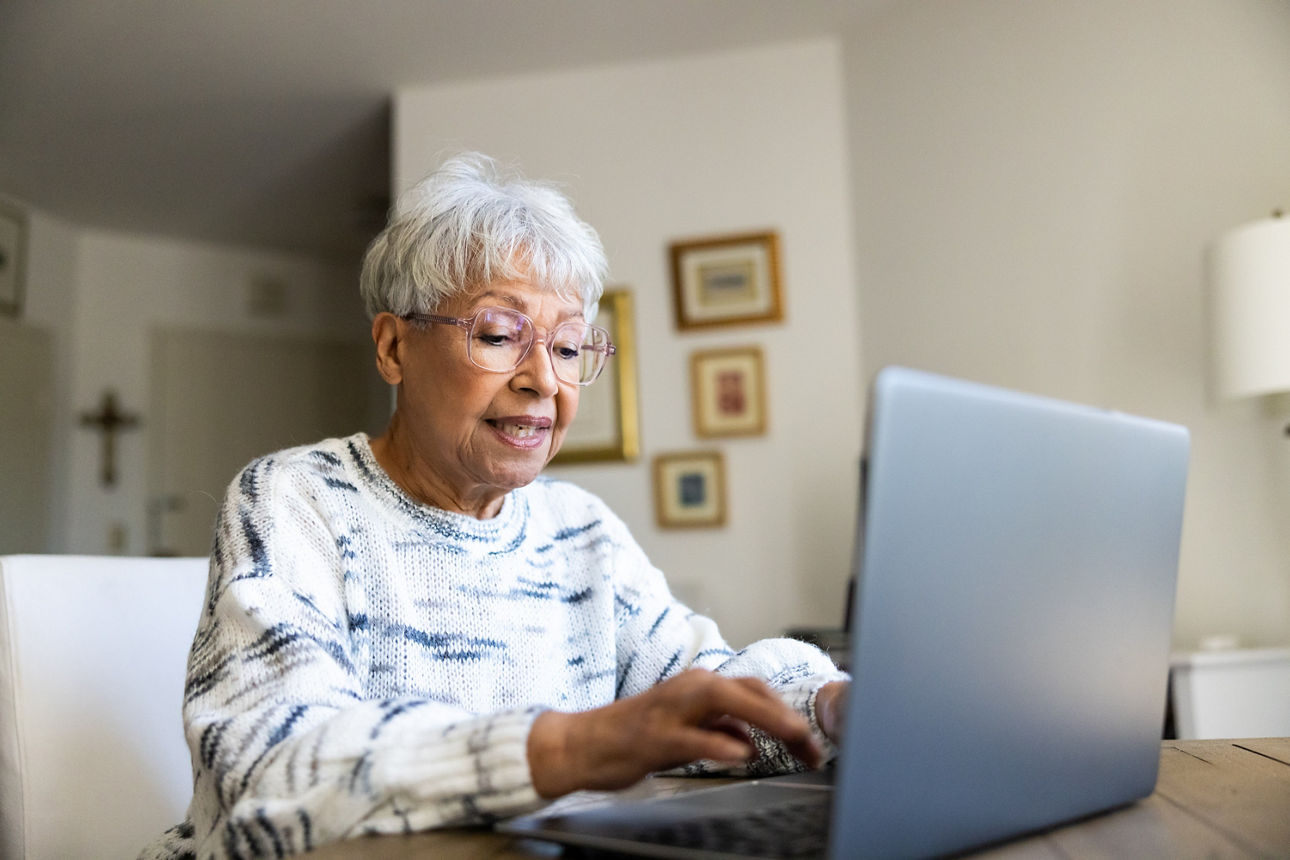 Eine ältere Frau sitzt zu Hause am Tisch und nutzt einen Laptop.