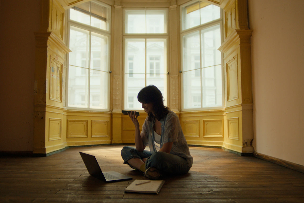 Eine junge Frau sitzt in einer leeren Wohnung vor einem Erker mit hohen Fenstern mit Laptop, Block und Stift auf dem Boden. Parallel telefoniert sie mit ihrem Handy via Lautsprecher. Eine junge Frau sitzt in einer leeren Wohnung vor einem Erker mit hohen Fenstern mit Laptop, Block und Stift auf dem Boden. Parallel telefoniert sie mit ihrem Handy via Lautsprecher.