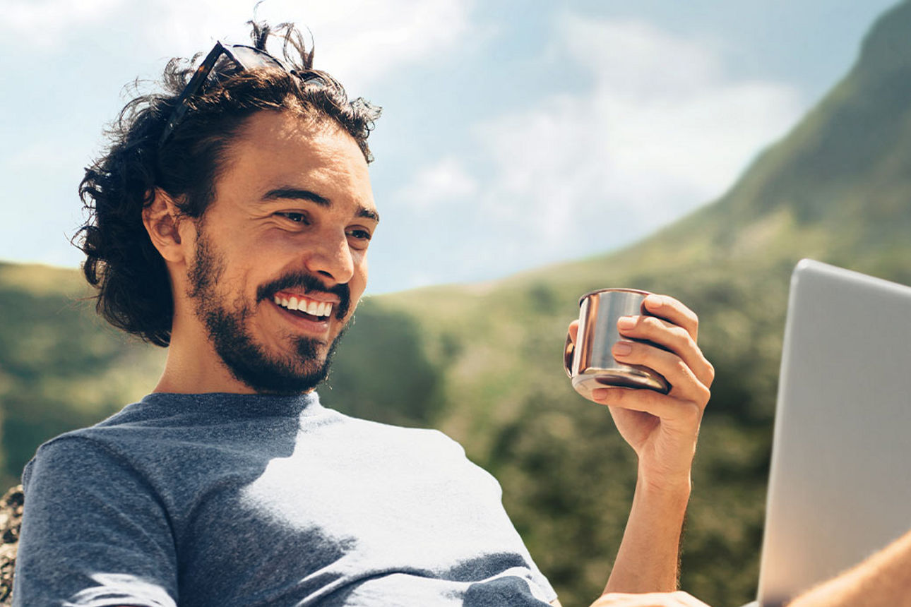 Bärtiger Mann sitzt vor Alpenpanorama lächelnd vor einem Laptop und hält eine Tasse in der Hand