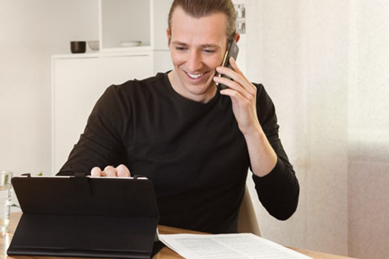 Ein Mann sitzt an einem Tisch und telefoniert, während er auf einem Tablet arbeitet.  