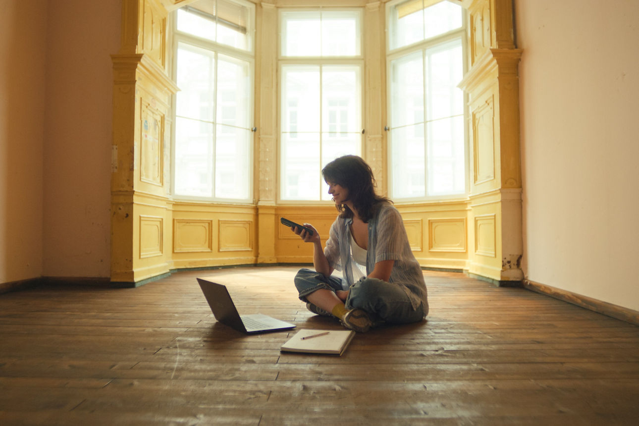 Eine junge Frau sitzt in einer leeren Wohnung vor einem Erker mit hohen Fenstern mit Laptop, Block und Stift auf dem Boden. Parallel tippt sie auf ihrem Handy.