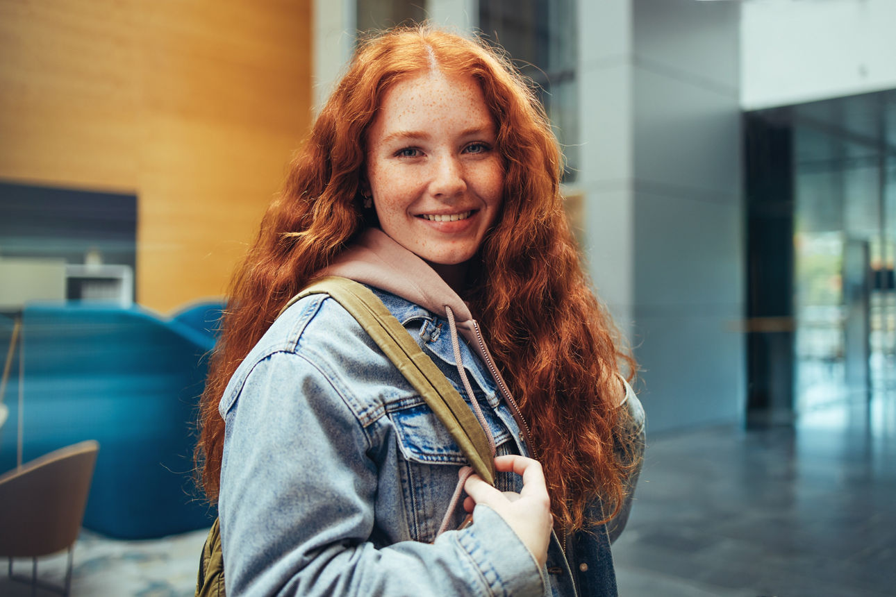 Eine junge, rothaarige Frau mit Umhängetasche lächelt in die Kamera