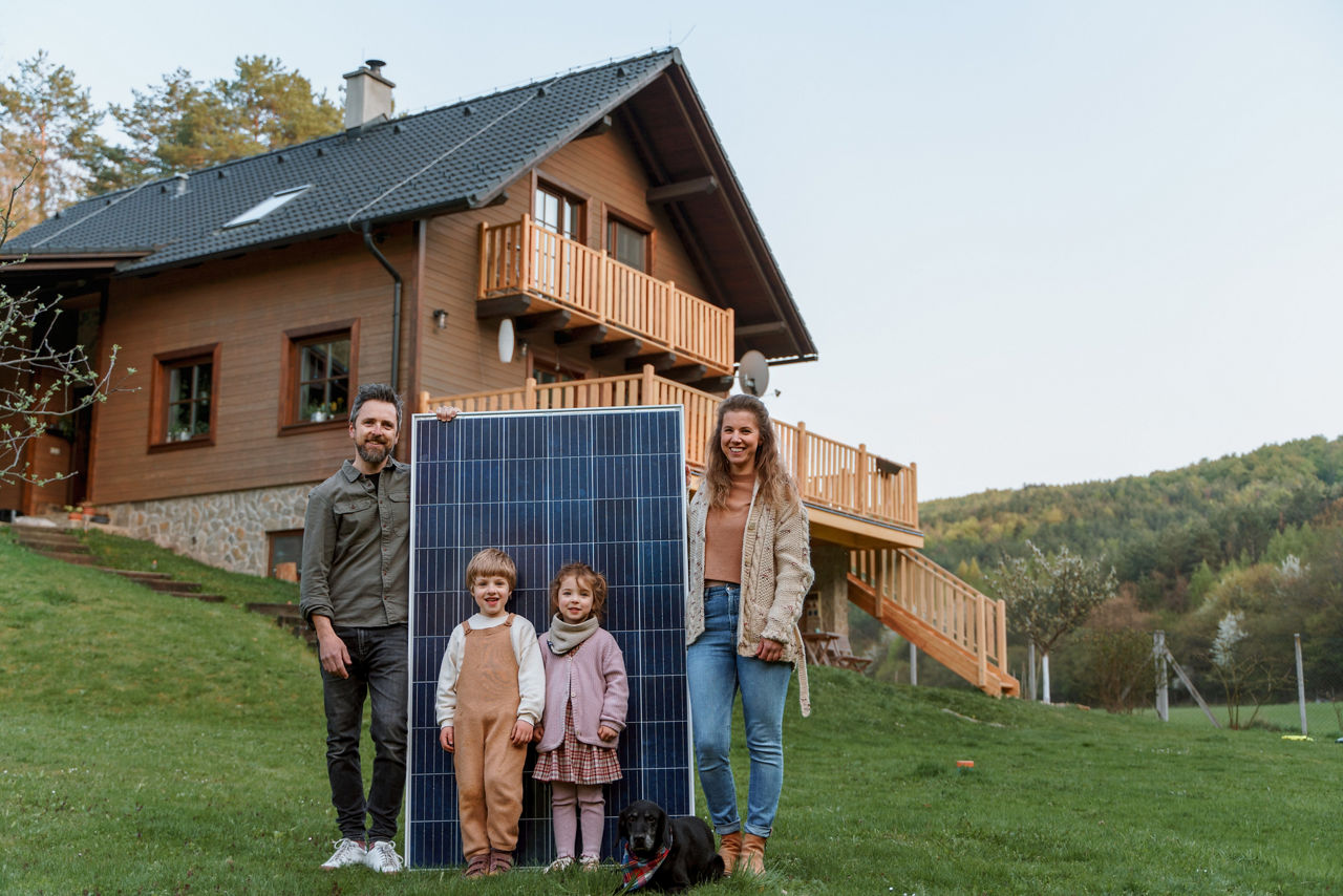 Eine Familie mit zwei Kindern & Hund steht mit einem Solarpanel vor einem Haus auf einer Wiese