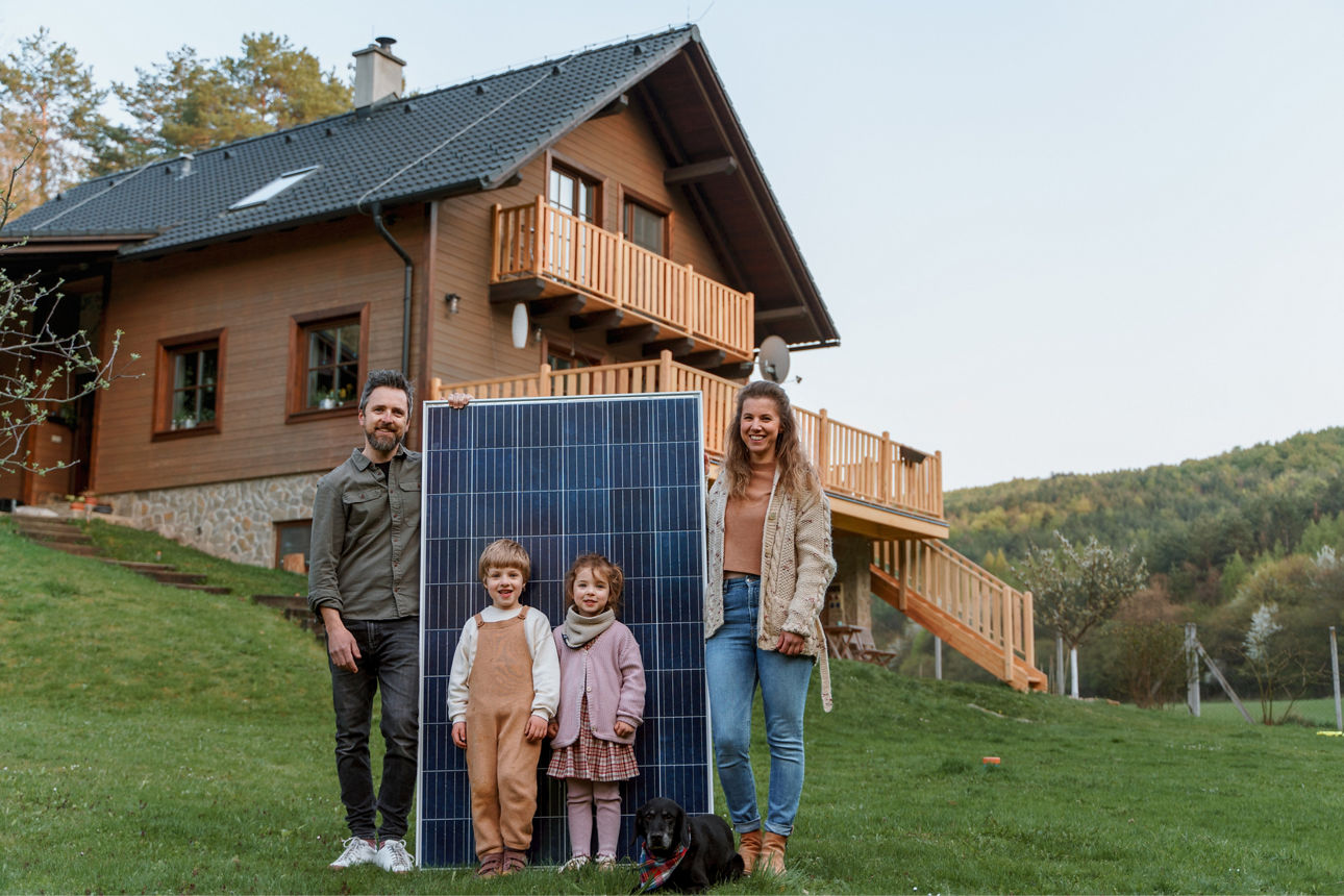 Eine Familie mit zwei Kindern & Hund steht mit einem Solarpanel vor einem Haus auf einer Wiese