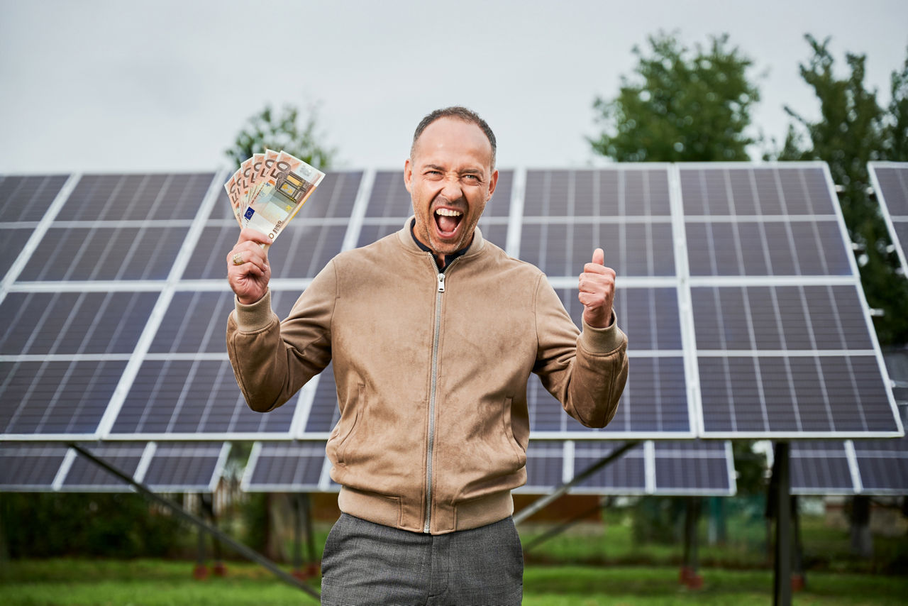 Ein Mann steht mit einer Handvoll Geldscheinen vor einer Solaranlage und jubelt sehr