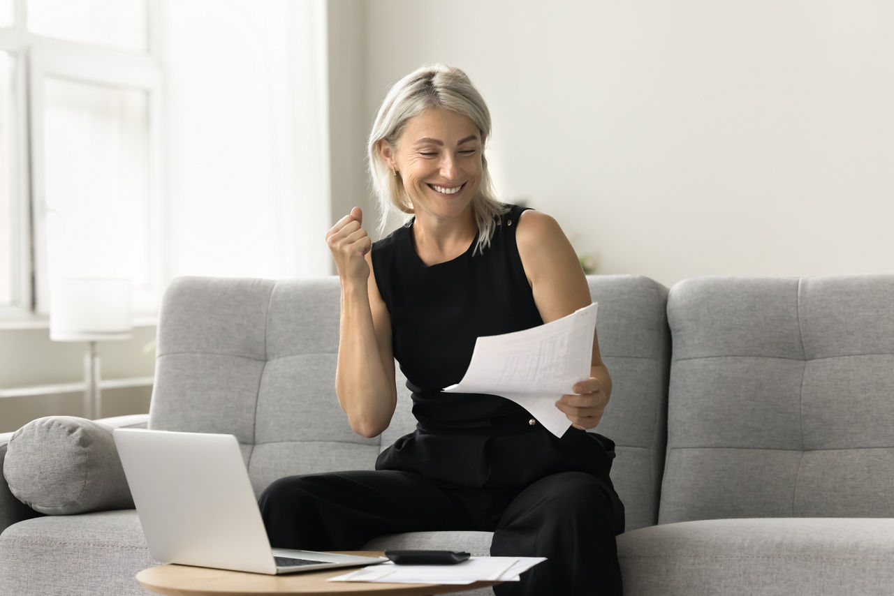Happy successful mature investor woman celebrating profit, income from investment, retirement savings, reading financial paper document, making winner hand, smiling, sitting at laptop at home