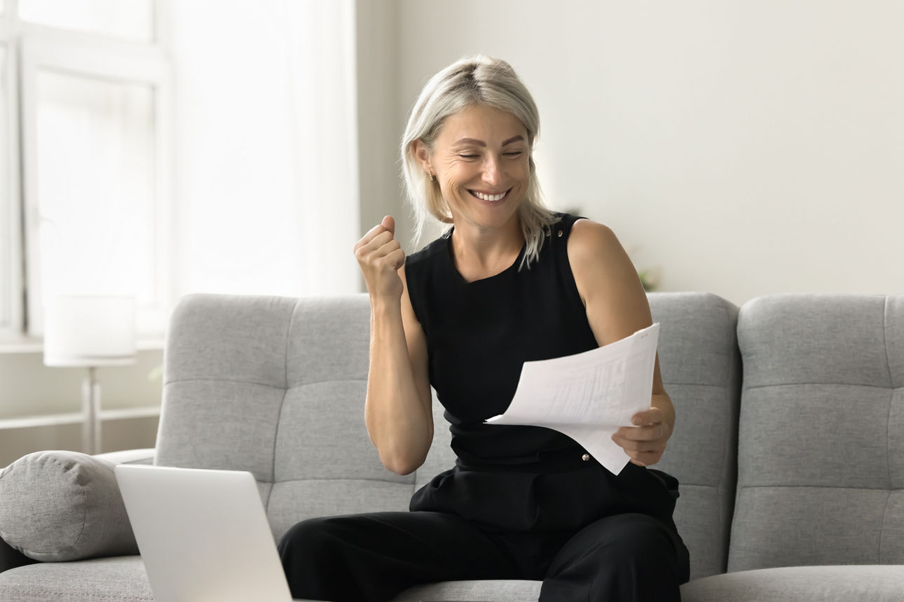 Happy successful mature investor woman celebrating profit, income from investment, retirement savings, reading financial paper document, making winner hand, smiling, sitting at laptop at home
