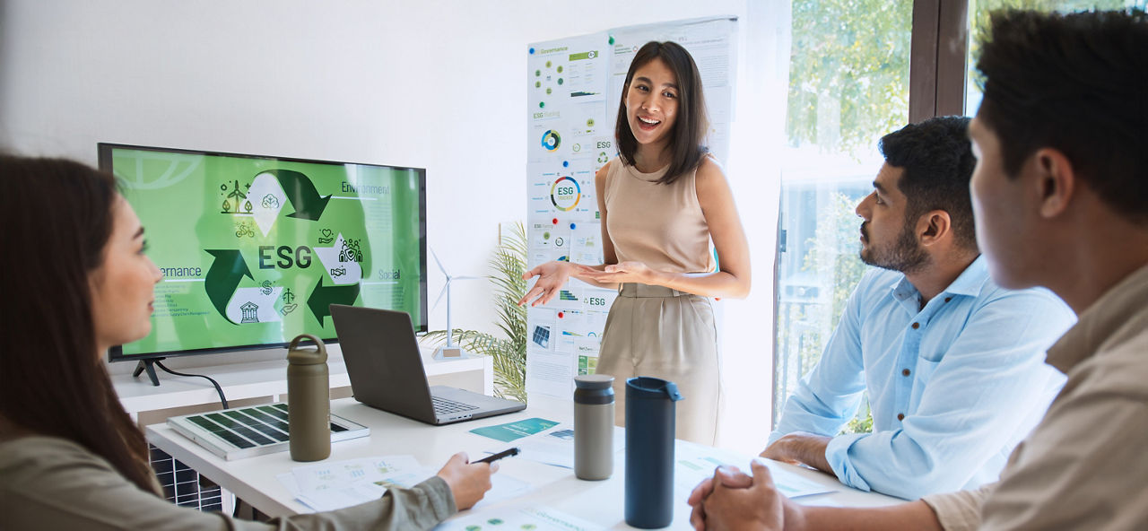 Vier Personen sitzen und stehen rund um einen Besprechungstisch und blicken auf einen Bildschirm mit den Worten "ESG"