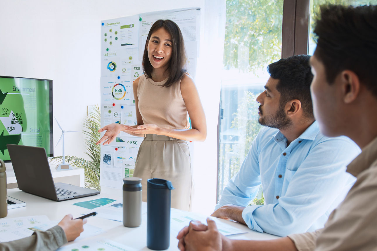 Vier Personen sitzen und stehen rund um einen Besprechungstisch und blicken auf einen Bildschirm mit den Worten "ESG"