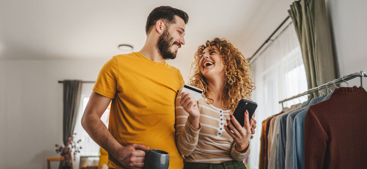 Ein Paar steht in Arm in Arm, er mit einer Tasse und sie mit EC-Karte und Handy