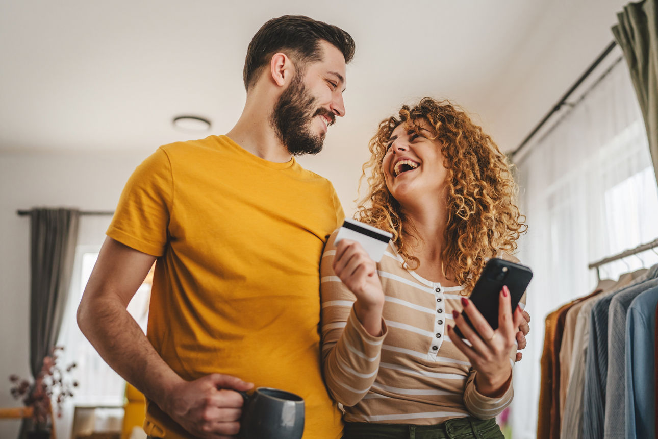 Ein Paar steht in Arm in Arm, er mit einer Tasse und sie mit EC-Karte und Handy