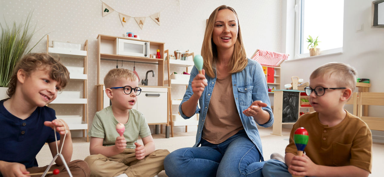 Eine Kindergärtnerin sitzt mit drei Kindern zusammen, sie spielen auf Rasseln und einer Triangel