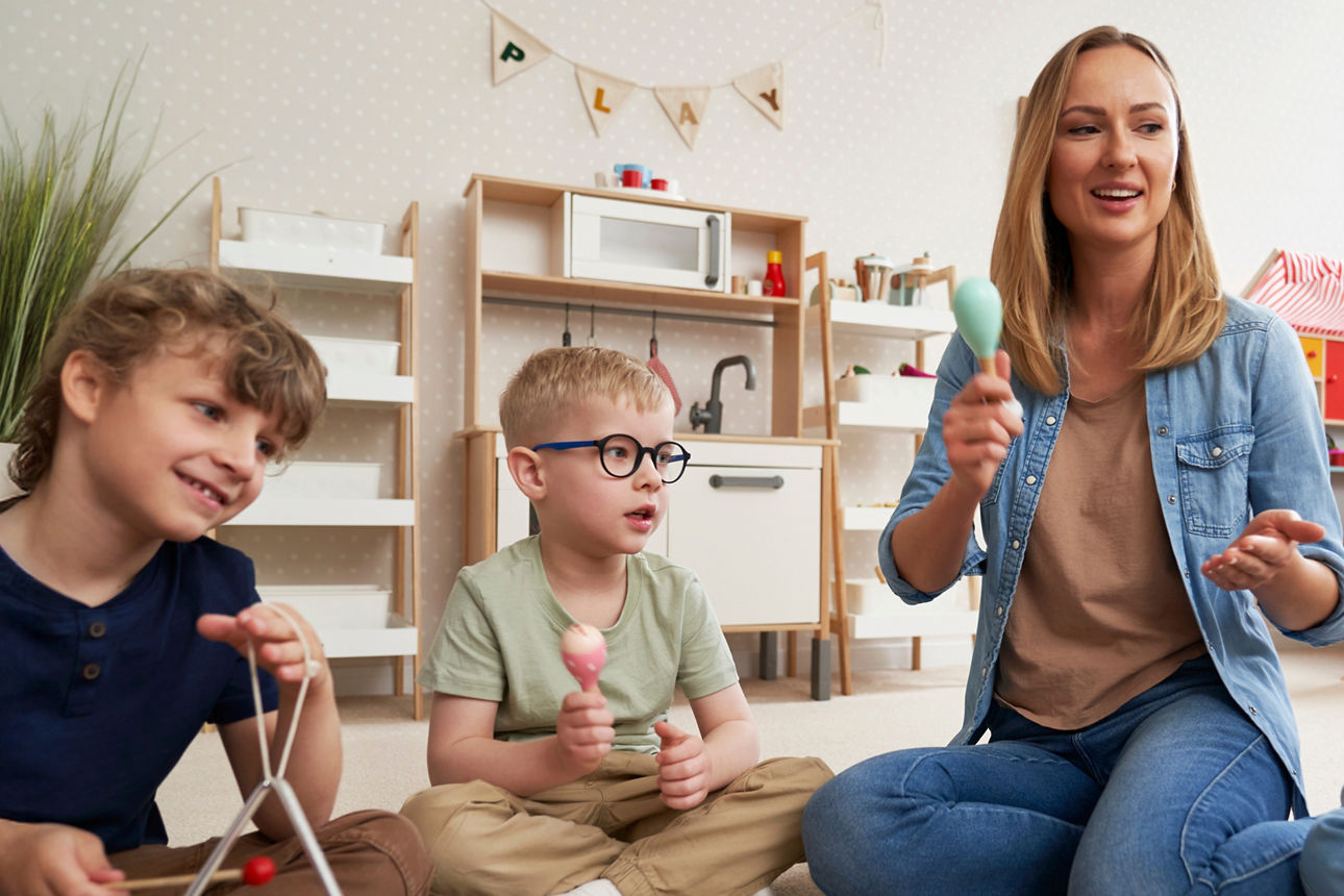 Eine Kindergärtnerin sitzt mit drei Kindern zusammen, sie spielen auf Rasseln und einer Triangel