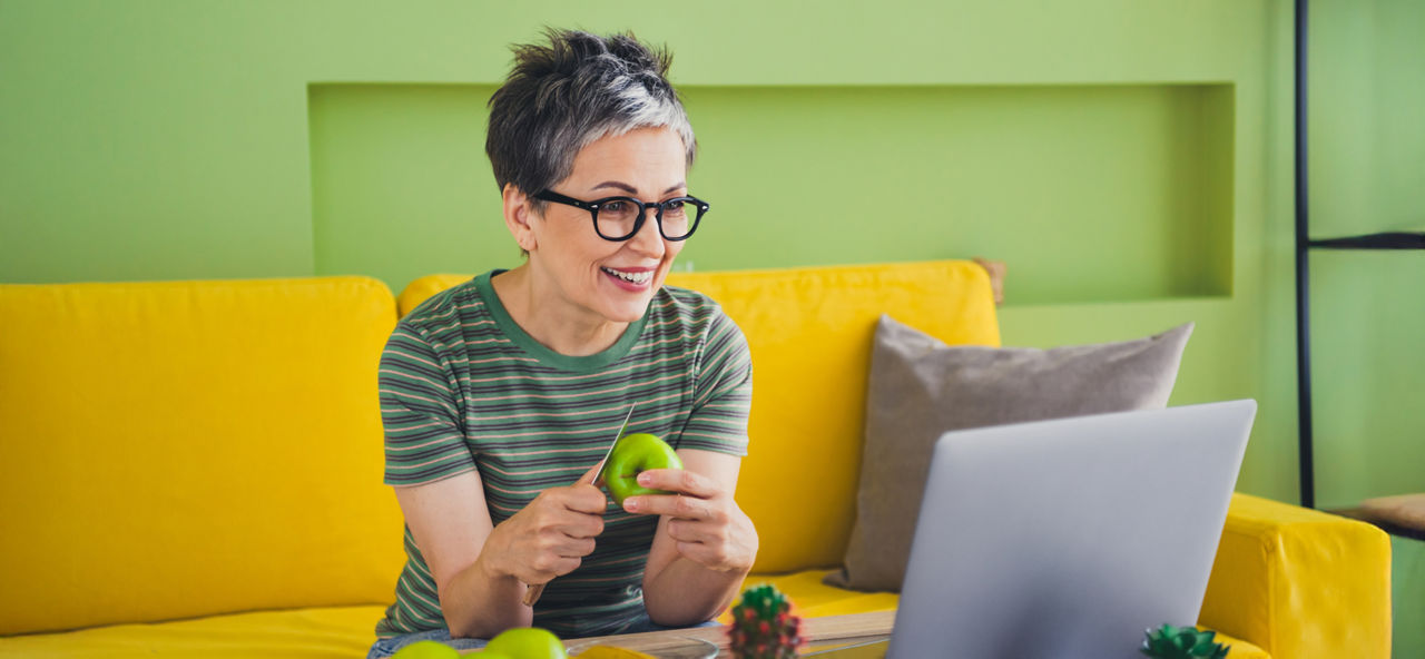 Eine Frau sitzt auf einer Couch, schält einen Apfel und blickt in einen Laptop