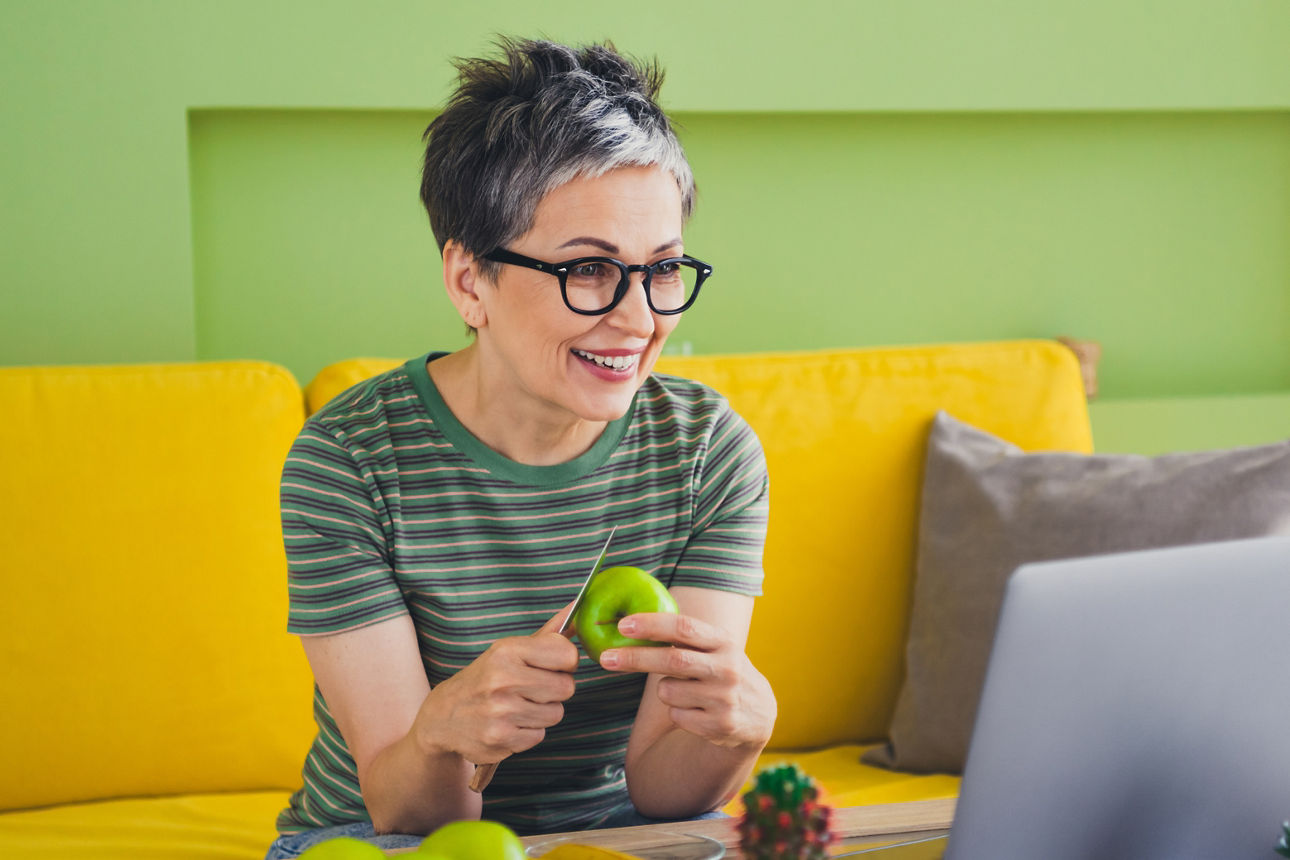 Eine Frau sitzt auf einer Couch, schält einen Apfel und blickt in einen Laptop