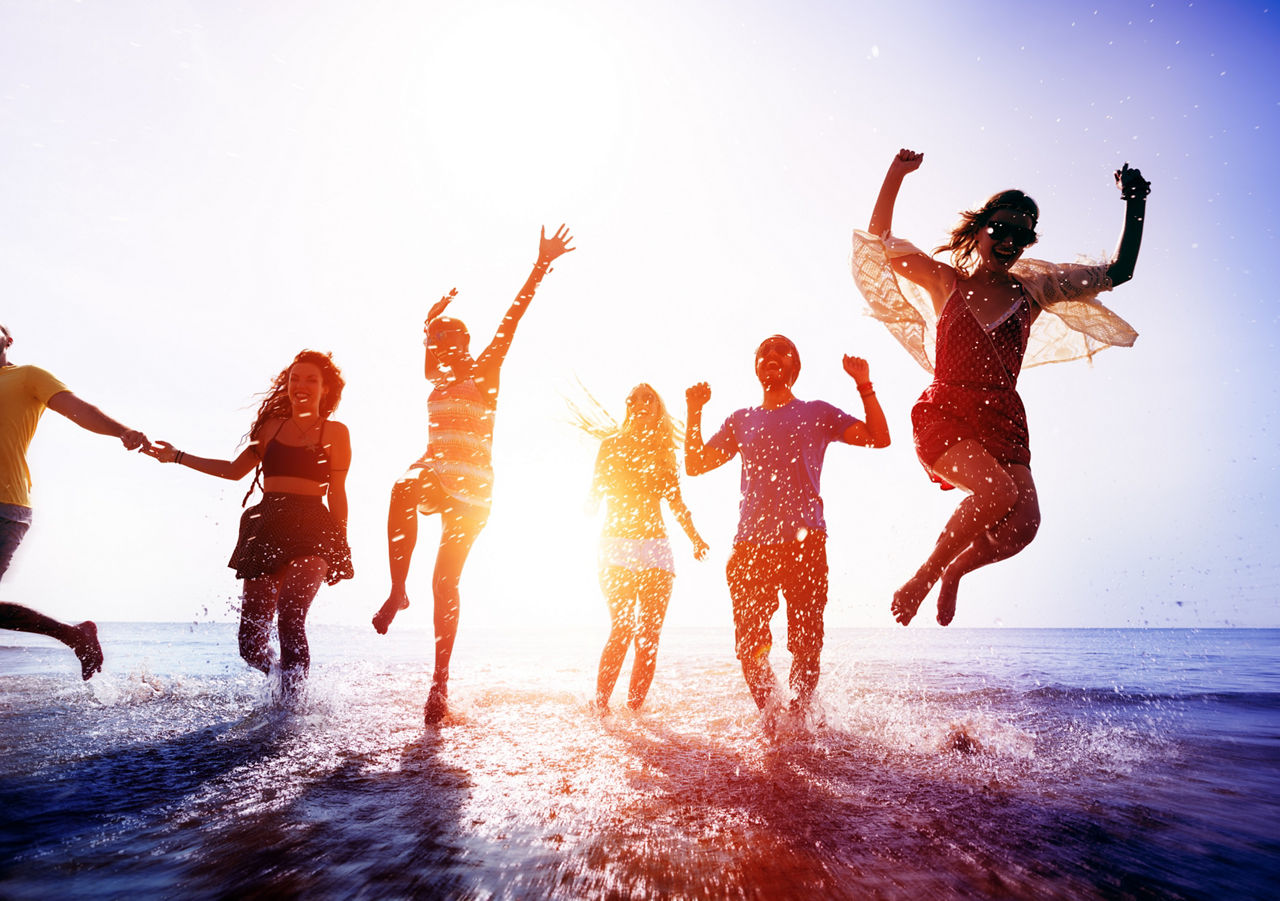 Eine Gruppe junger Menschen läuft fröhlich springend am Strand, im Hintergrund das Meer und die Sonne
