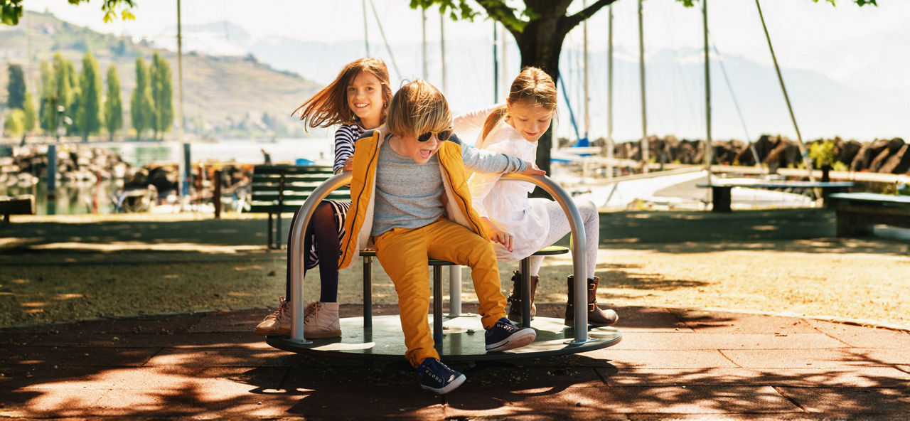Drei Kinder sitzen auf einem Karusell im Freien vor einem See