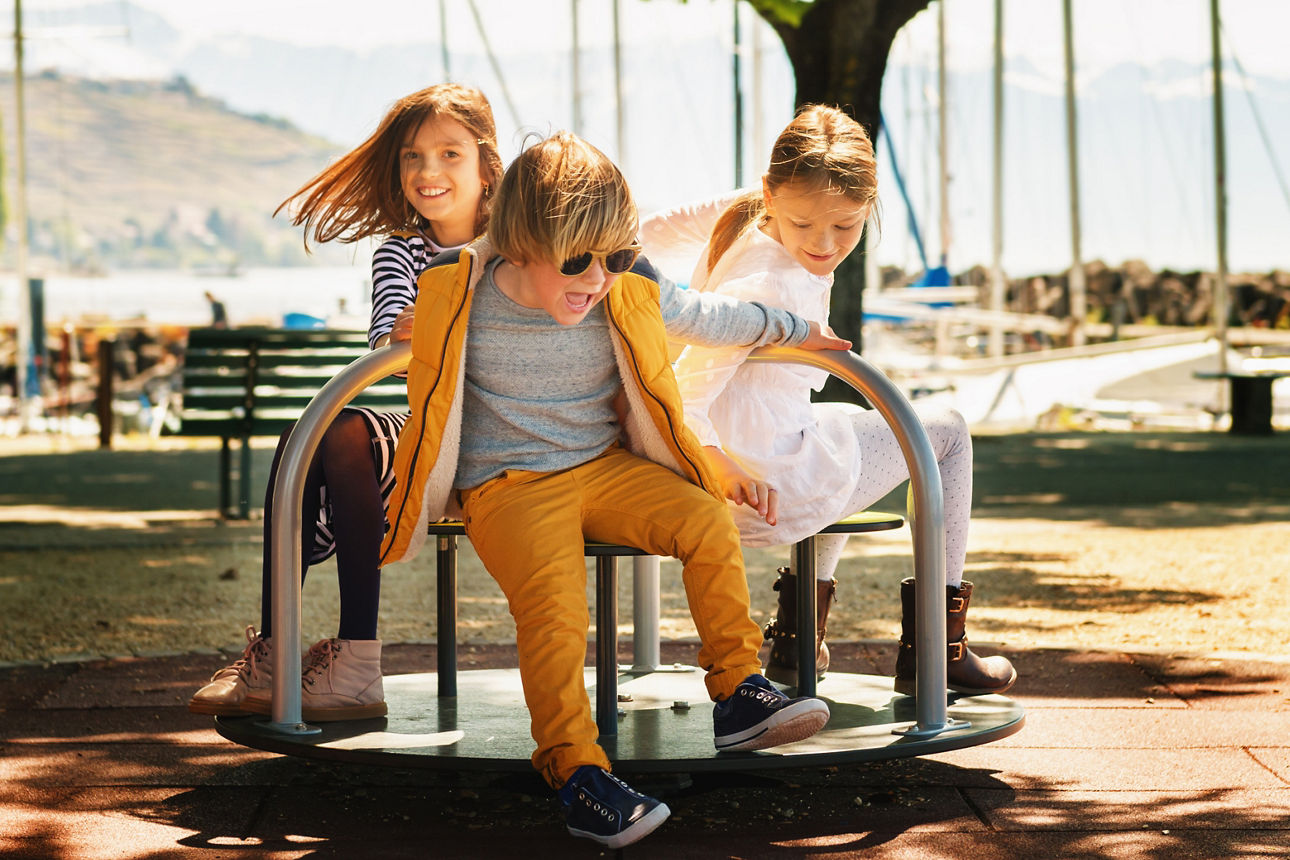 Drei Kinder sitzen auf einem Karusell im Freien vor einem See
