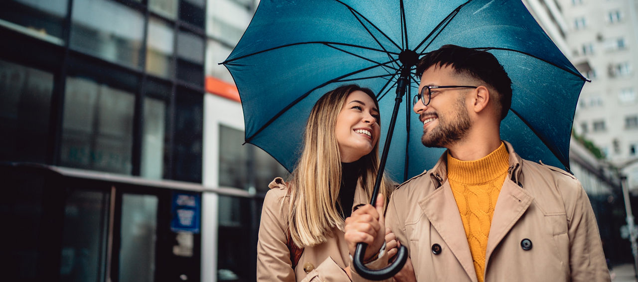 Ein schönes lächelndes junges Touristenpaar, das mit einem Koffer und einem großen Regenschirm im Regen spazieren geht