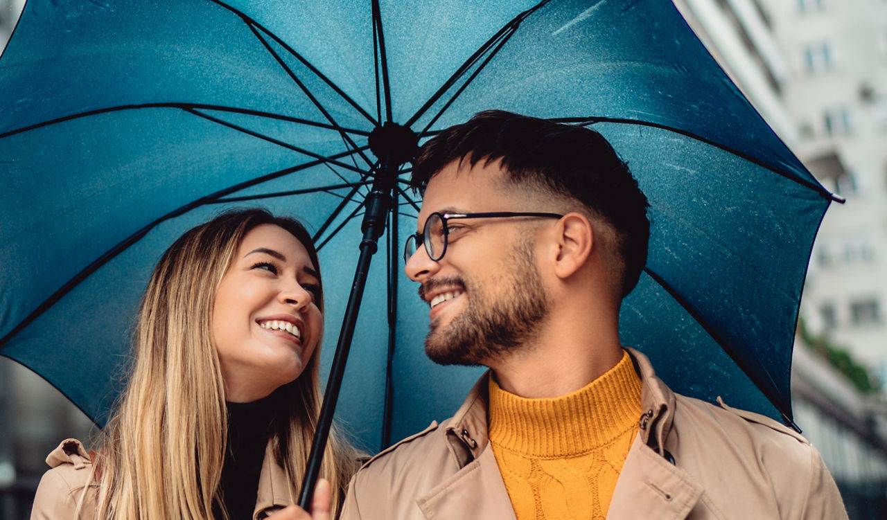 Ein hübsches, lächelndes junges Paar von Touristen geht im Regen mit Koffer und großem Schirm spazieren.