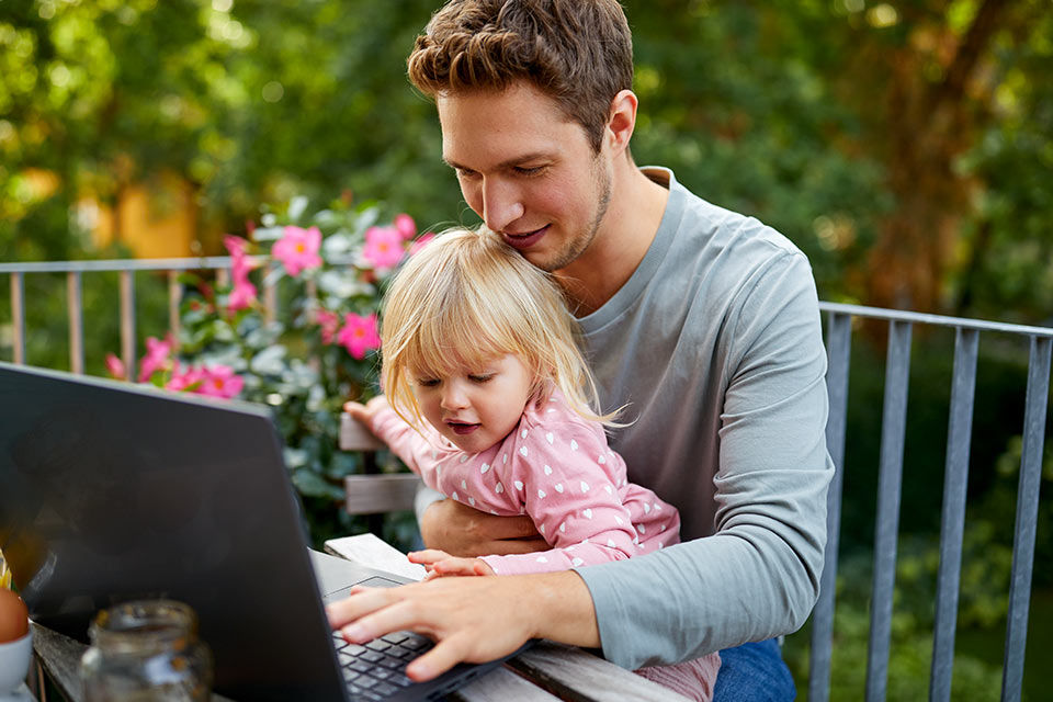 Mann sitzt mit Tochter auf Balkon an einem Laptop