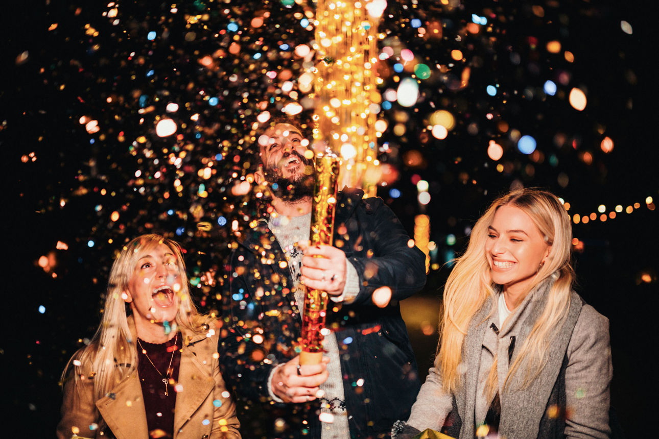 Drei Personen feiern mit einer sprühenden Konfettikanone in der Hand vor dunklem Hintergrund.