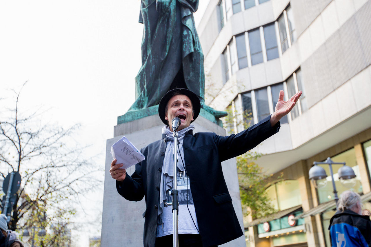 Ein Mann mit Zylinder steht draussen vor einem Mikrofon und redet.