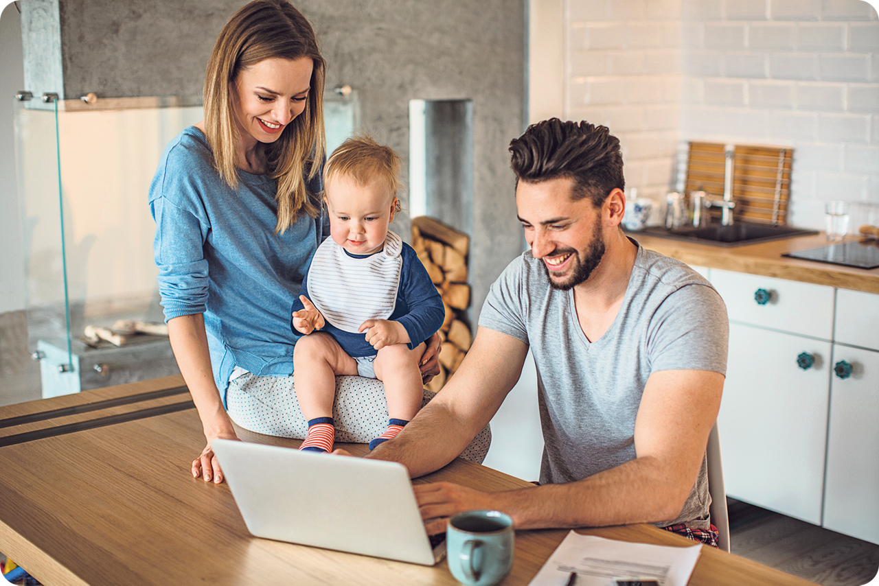 Ein Mann sitzt am Küchentisch und schaut lächelnd auf sein Notebook. Eine Frau mit einem Baby auf dem Schoß sitzt lächelnd daneben.