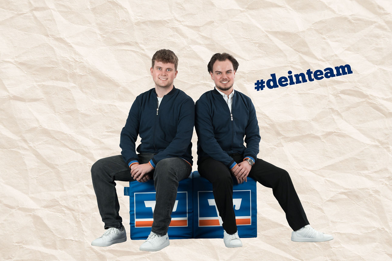 Zwei Männer sitzen Rücken an Rücken auf blauen VR-Bank-Sitzwürfeln vor hellem Hintergrund.