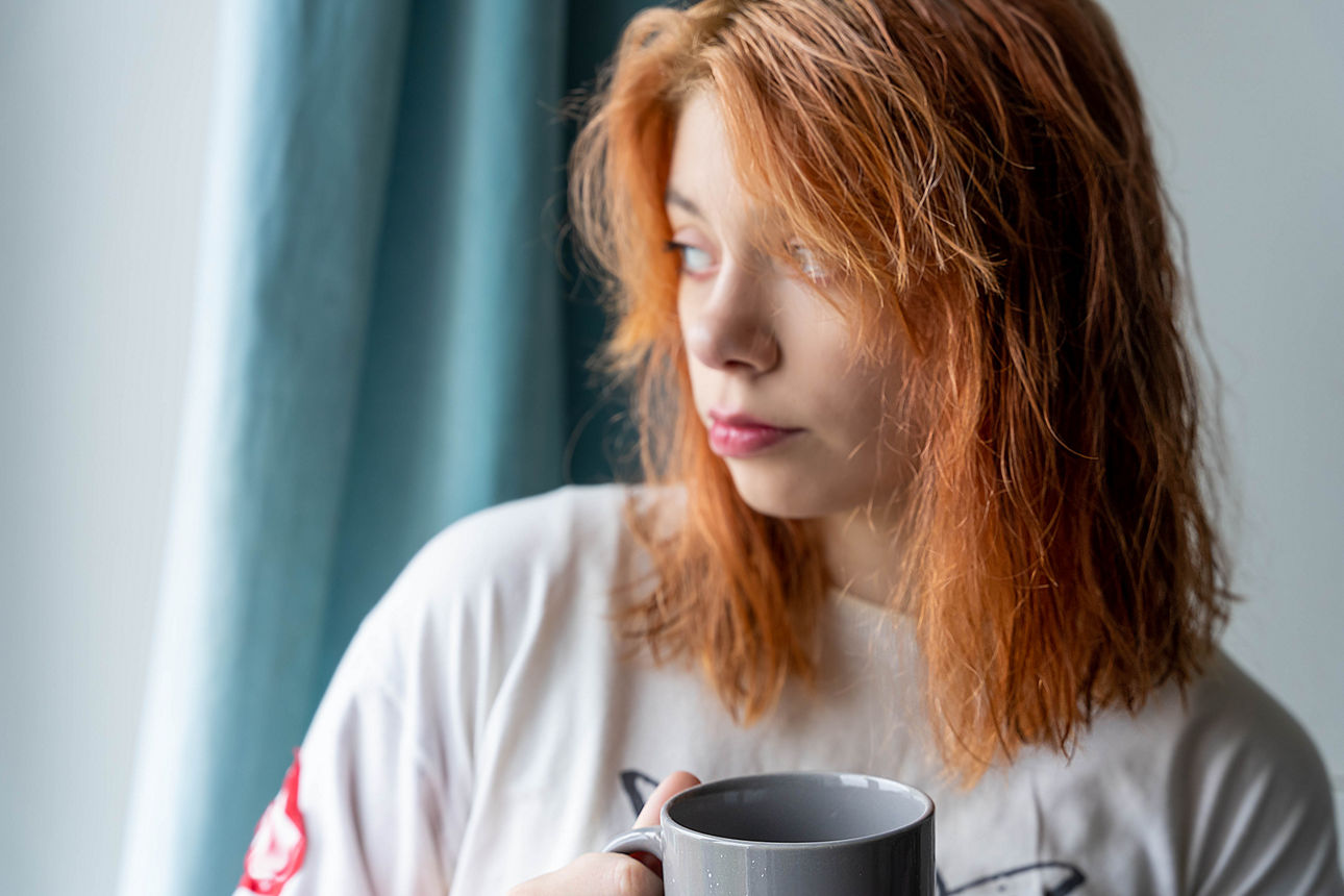 Person mit rotem Haar hält eine Tasse und blickt aus dem Fenster.