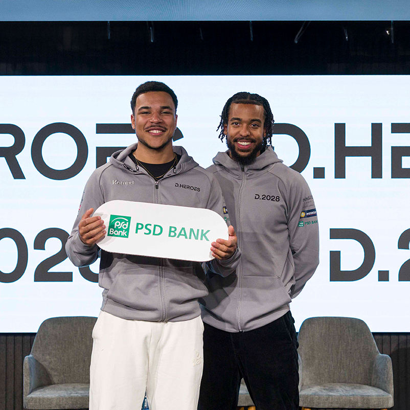 Zwei Sportler des Düsseldorfer Olympiateams halten ein Schild mit dem Logo der PSD Bank hoch.