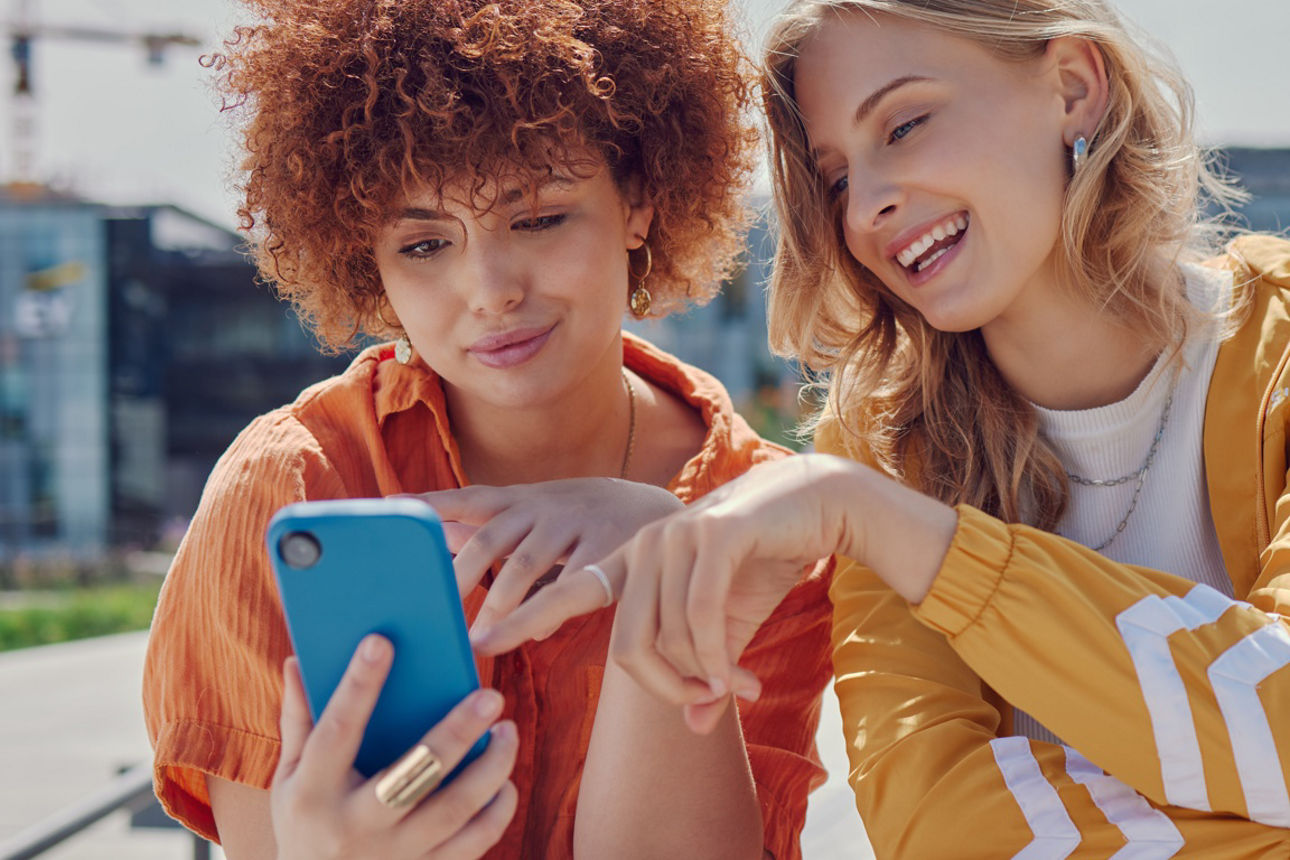 Zwei fröhliche junge Frauen sitzen in städtischer Kulisse und blicken auf ein Smartphone