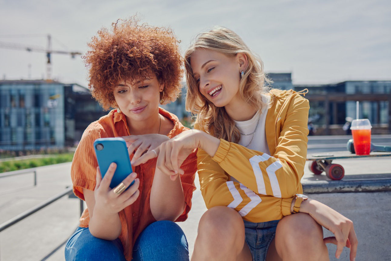 Zwei fröhliche junge Frauen sitzen in städtischer Kulisse und blicken auf ein Smartphone