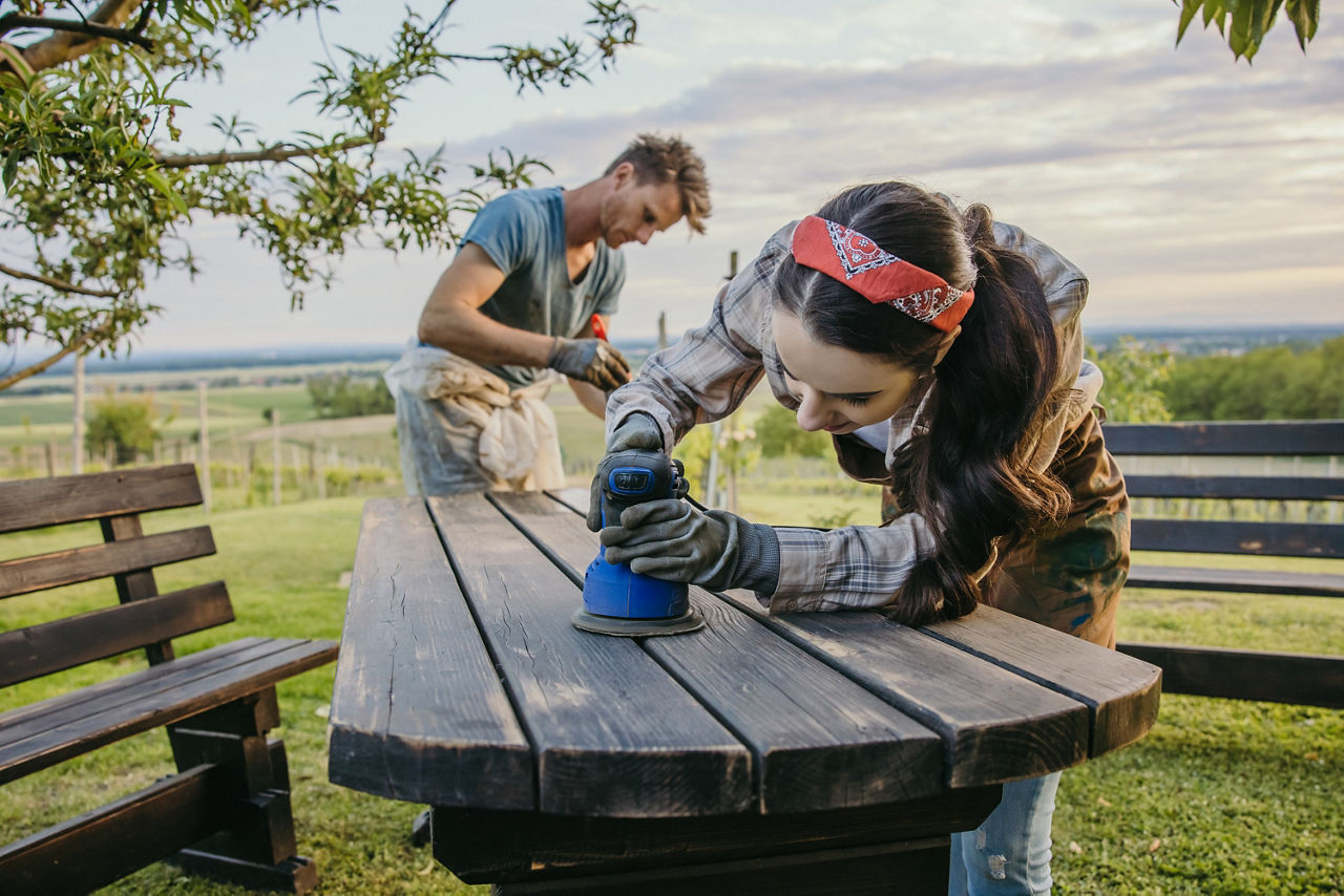 Im Freien bei schönem Wetter arbeiten eine Frau im Vordergrund und ein Mann im Hintergrund an einem Holztisch. Sie schleifen und streichen diesen. 