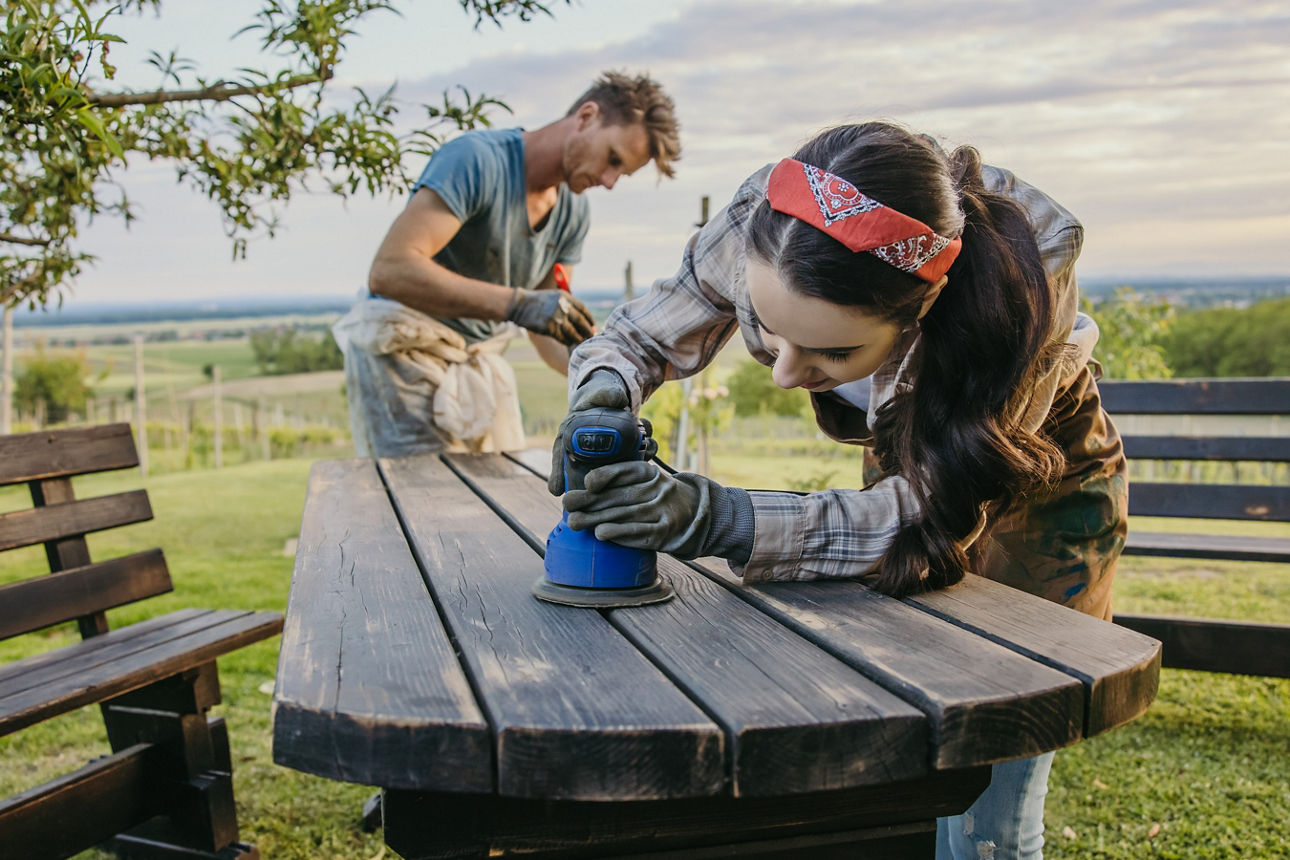 Im Freien bei schönem Wetter arbeiten eine Frau im Vordergrund und ein Mann im Hintergrund an einem Holztisch. Sie schleifen und streichen diesen. 