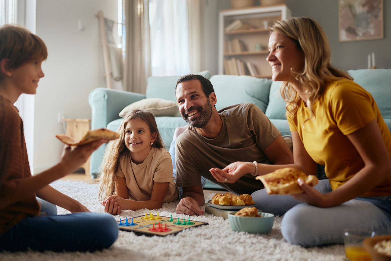 Eine vierköpfige Familie spielt auf dem Boden ein Brettspiel und isst nebenbei.