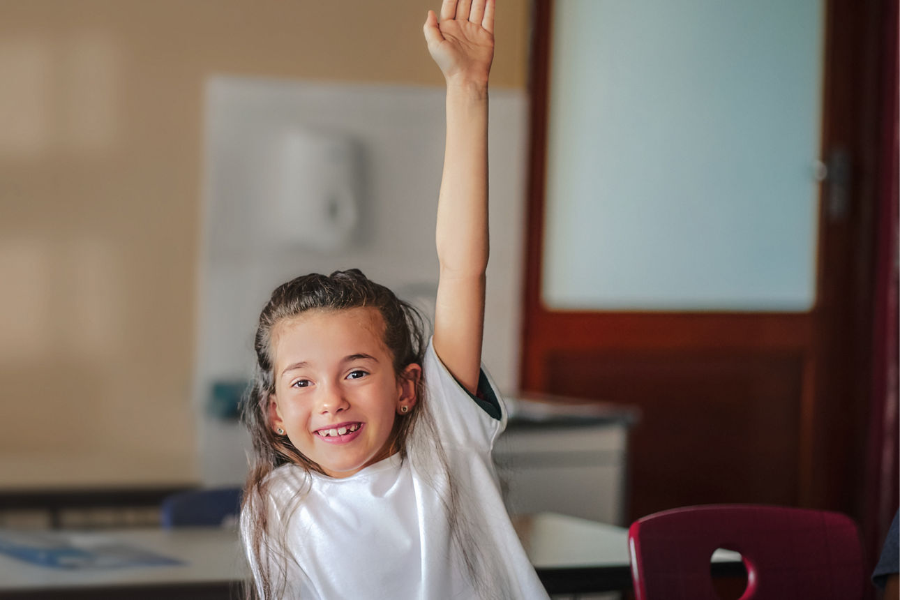 Ein Mädchen sitzt im Klassenzimmer und meldet sich mit erhobener Hand.