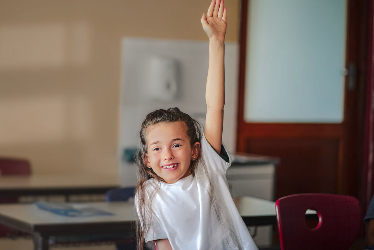 Ein Mädchen sitzt im Klassenzimmer und meldet sich mit erhobener Hand.