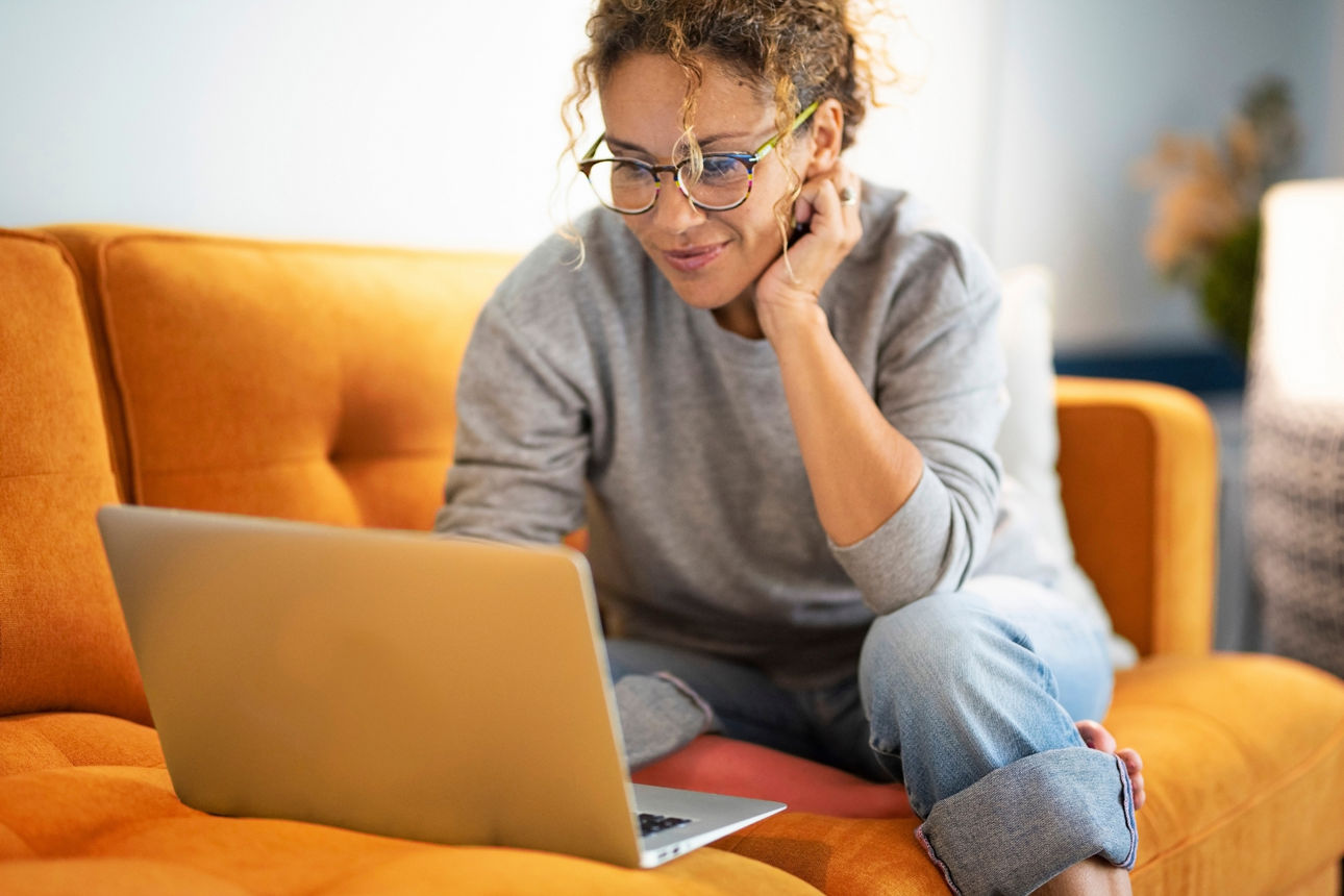 Smiling woman in vibrant orange top and denim jeans sitting comfortably on couch, deeply engaged with her smartphone in cozy home interior