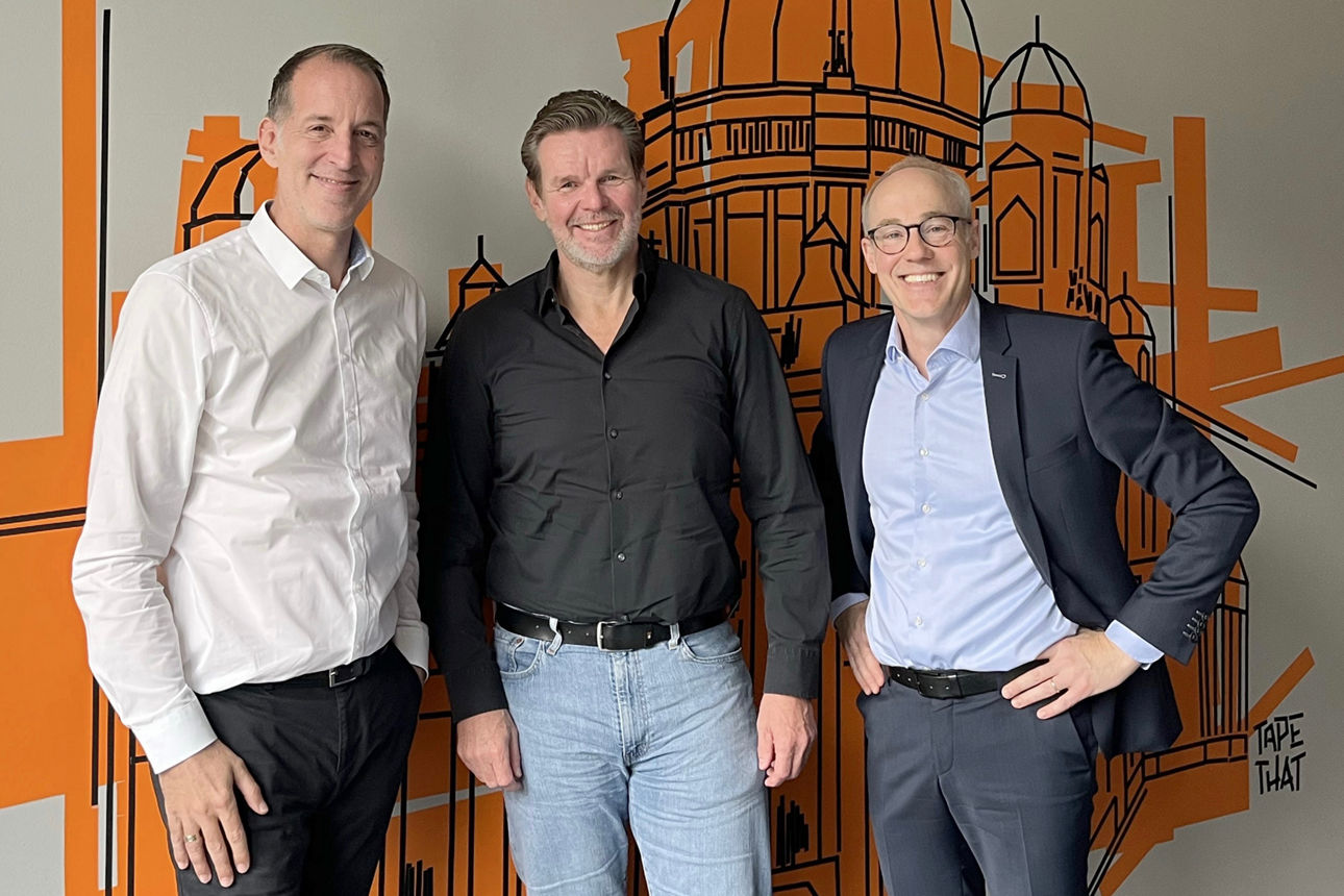 Drei Männer stehen nebeneinander vor einer Wand und lächeln in die Kamera