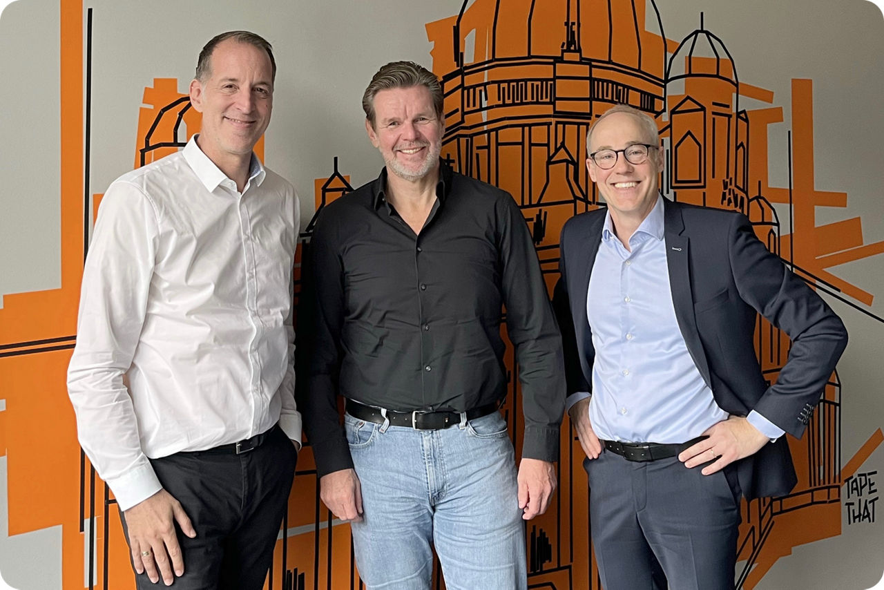 Drei Männer stehen nebeneinander vor einer Wand und lächeln in die Kamera