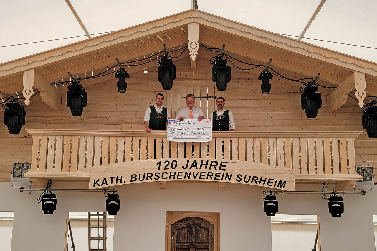 Drei Männer stehen auf dem Holzbalkon und halten einen übergroßen symbolischen Bar-Scheck in der Hand. 