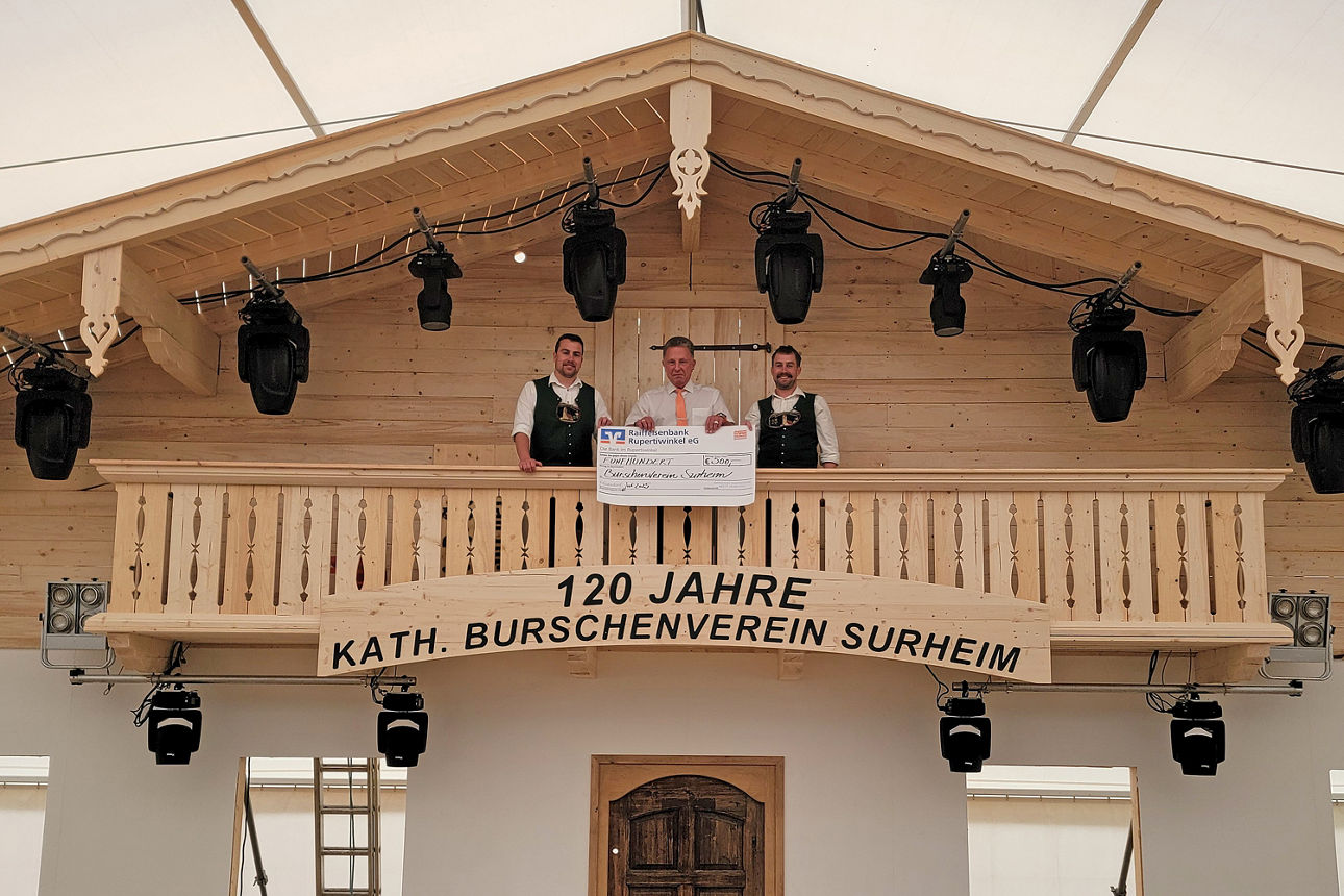 Drei Männer stehen auf dem Holzbalkon und halten einen übergroßen symbolischen Bar-Scheck in der Hand. 