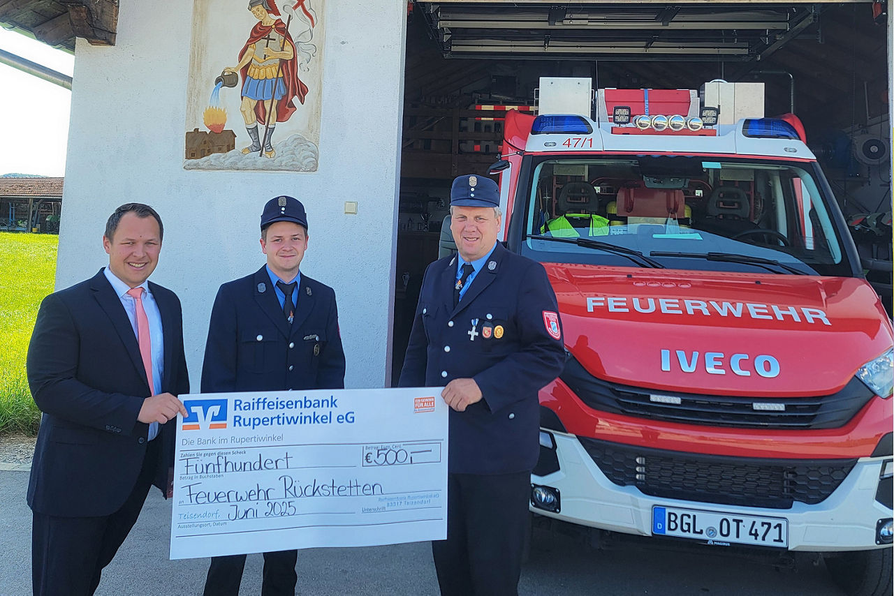 Drei Männer stehen vor einem Feuerwehrauto und halten einen übergroßen symbolischen Bar-Scheck hoch.