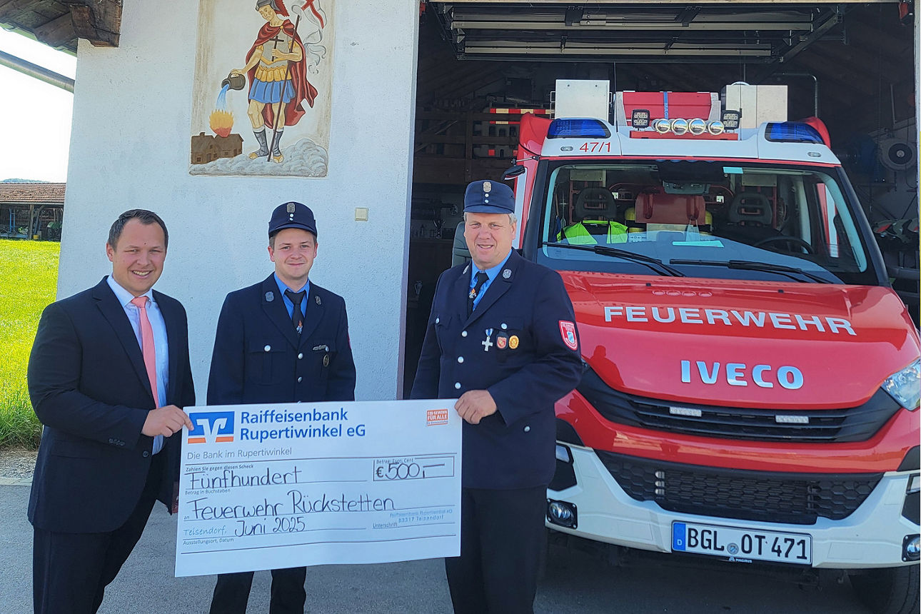 Drei Männer stehen vor einem Feuerwehrauto und halten einen übergroßen symbolischen Bar-Scheck hoch.