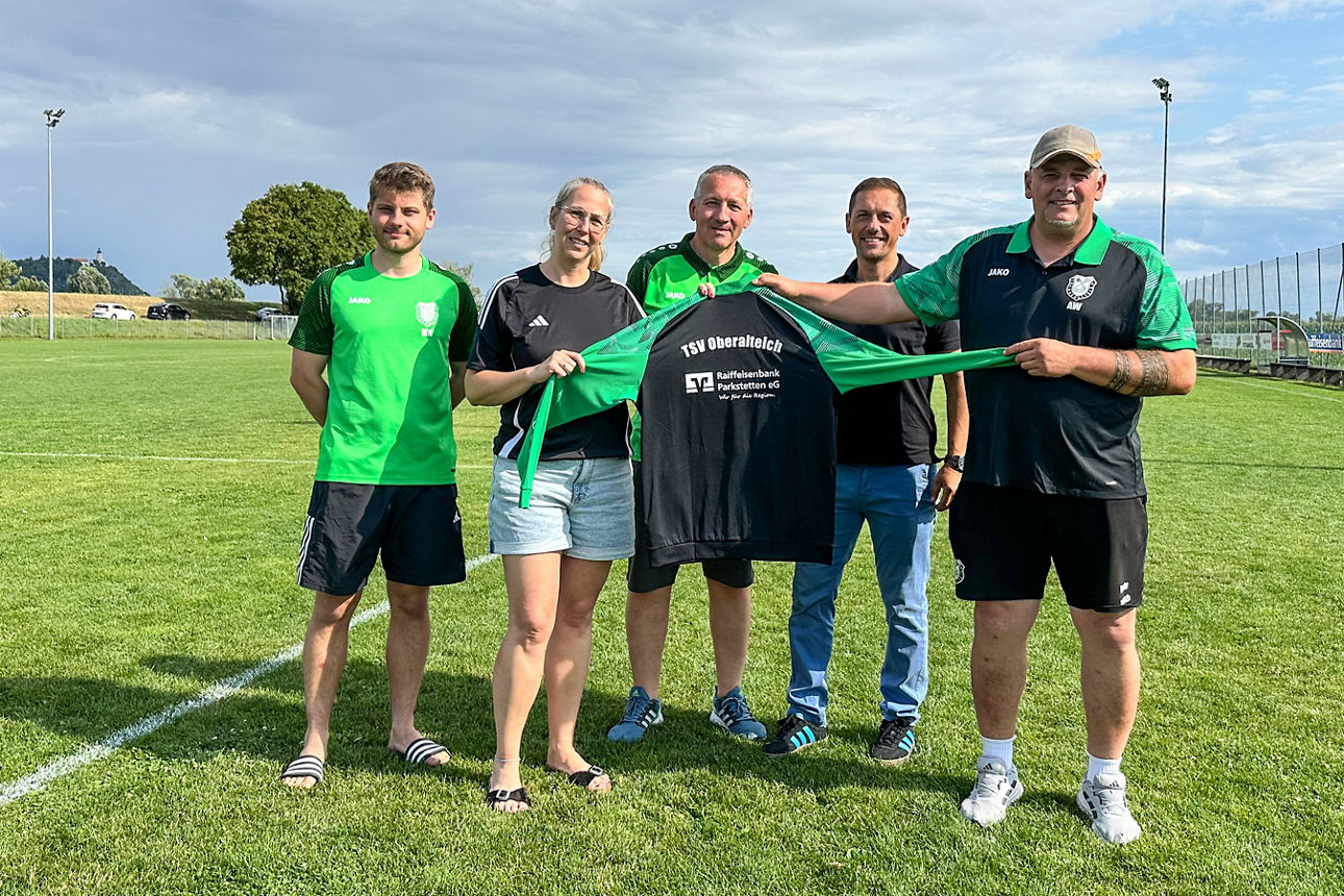 Fünf Personen auf Sportplatz präsentieren Trikot, gemeinsames Foto bei Trikotübergabe im Freien.