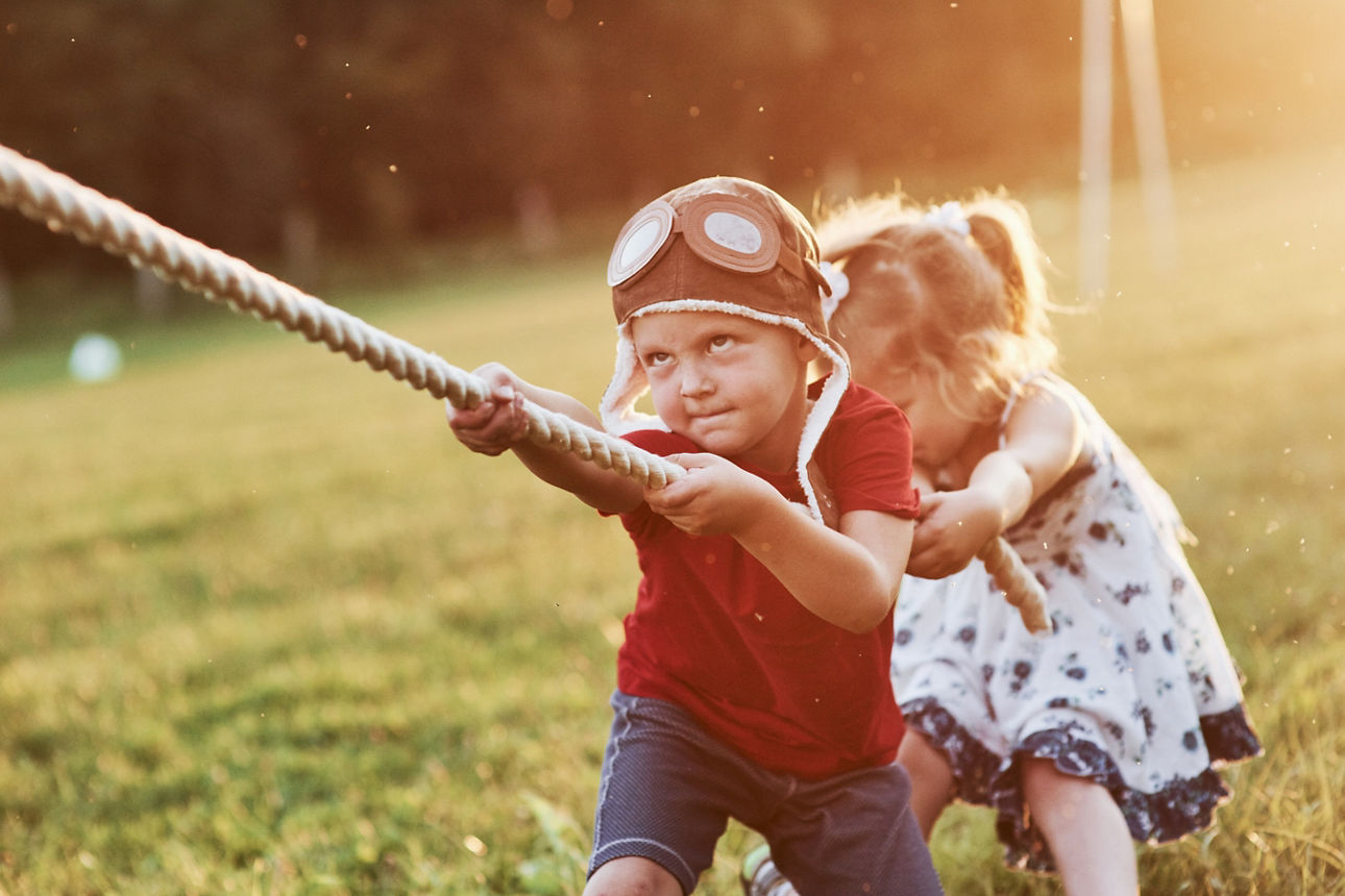 Zwei Kinder, ein Junge im Vordergrund und ein Mädchen dahinter, ziehen an einem Seil. Sie sind dabei draußen und es ist schönes Wetter.